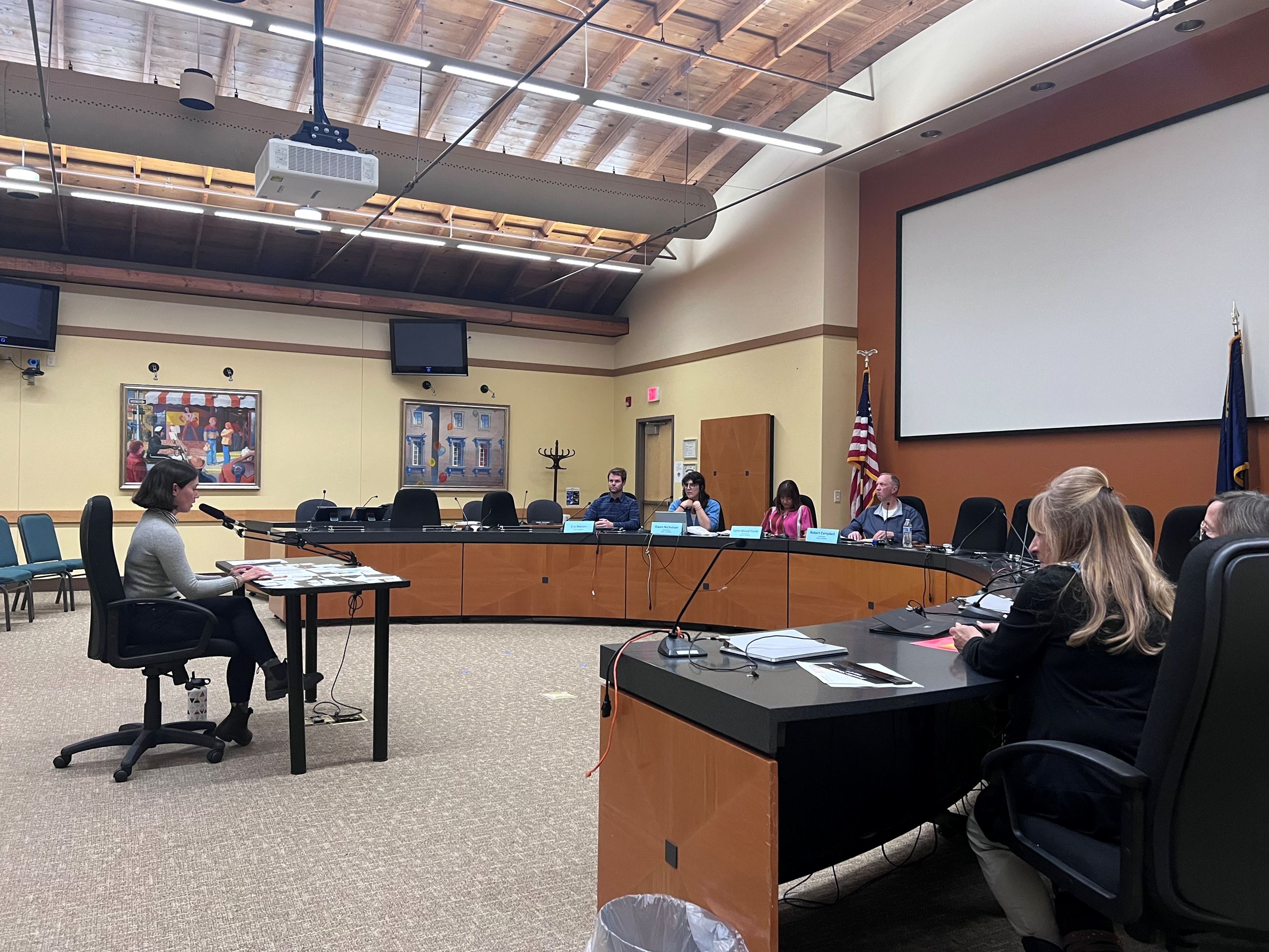 A woman at a desk in city council chambers with people gathered around the horseshoe table